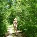 An unclothed woman in a sunhat walks on a path through an evergreen forest.