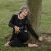 A grey haired woman stretches in a yoga pose at the foot of a tree.